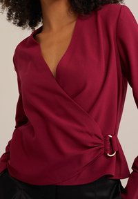 Blusa avvolgente color bordeaux in tessuto liscio con scollo a V profondo e dettaglio arricciato sul lato con anello decorativo dorato. Maniche lunghe aderenti.