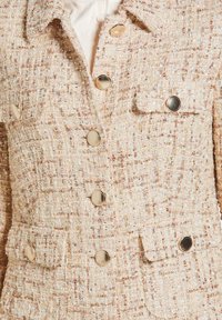 Beige tweedjasje met een gestructureerd oppervlak, voorzien van gouden knopen, een kraag en twee voorzakken.