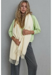 Écharpe blanche en tricot avec un motif en losange texturé et de longues franges, drapée sur un haut vert clair, sur un fond neutre.