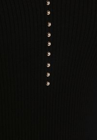 Top en tissu côtelé noir avec une rangée verticale de six boutons métalliques, ajoutant un détail texturé à la conception.