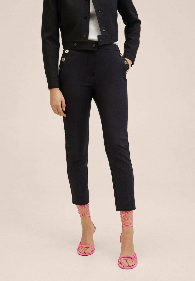 Femme portant un pantalon noir slim avec des boutons dorés, une veste noire courte, un haut blanc et des sandales à talons rose à lanières, debout avec une main dans la poche.