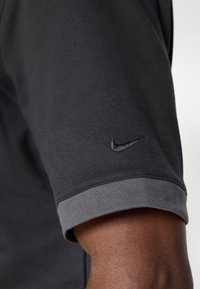 Cămizeta sport neagră cu detalii gri la mansete, având un logo Nike subtil, de culoare neagră, pe mânecă. Textură de material neted. Design cu mâneci scurte.