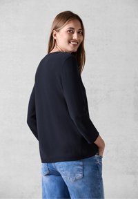 Maglione lungo a maniche lunghe di colore blu navy, con scollatura rotonda. Abbinato a jeans azzurri, che mostrano una vestibilità comoda e un design semplice.