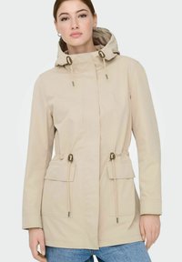Veste imperméable beige avec capuche, taille à cordon de serrage et deux poches avant. Texture douce et design épuré, agrémenté d'accents métalliques argentés.