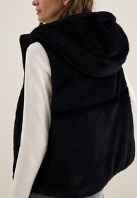 Svart pälsväst i fuskpäls med rymlig huva, som har en mjuk textur och dragkedja. Bärs över en ljus långärmad topp, vilket ger en avslappnad passform.