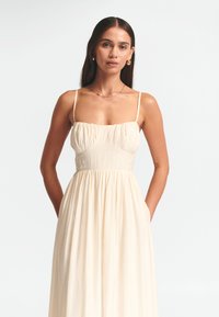 Vestido sin mangas de color crema, con un corsé ajustado y una falda fruncida. Tela texturada con un brillo sutil y bolsillos laterales.