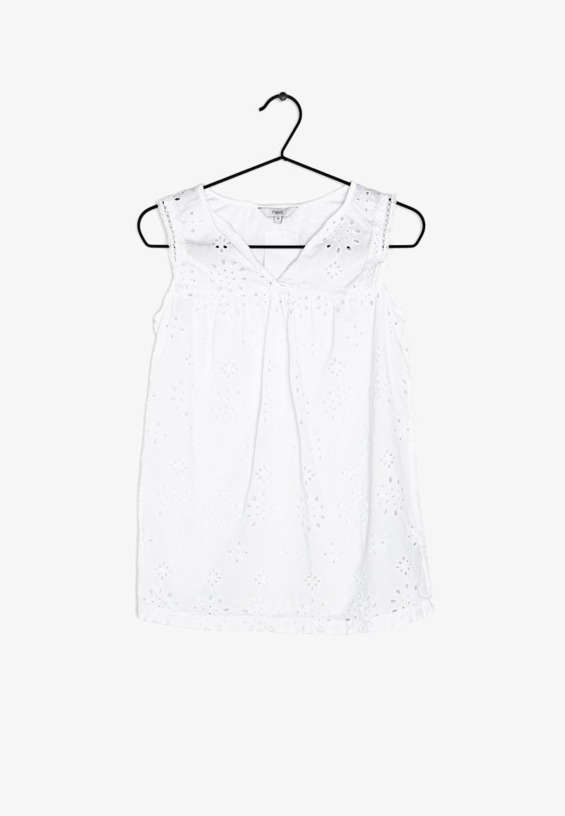 Blouse sans manches blanche avec broderies florales et accents en dentelle, présentant un décolleté en V et des détails froncés à la taille. Accrochée sur un crochet noir.