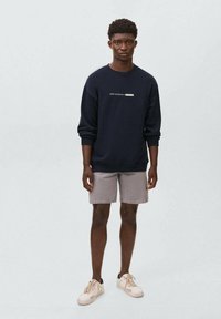 Felpa blu marina con testo, abbinata a pantaloni corti grigi chiari e sneaker beige. La felpa presenta un collo rotondo e polsini a costine.