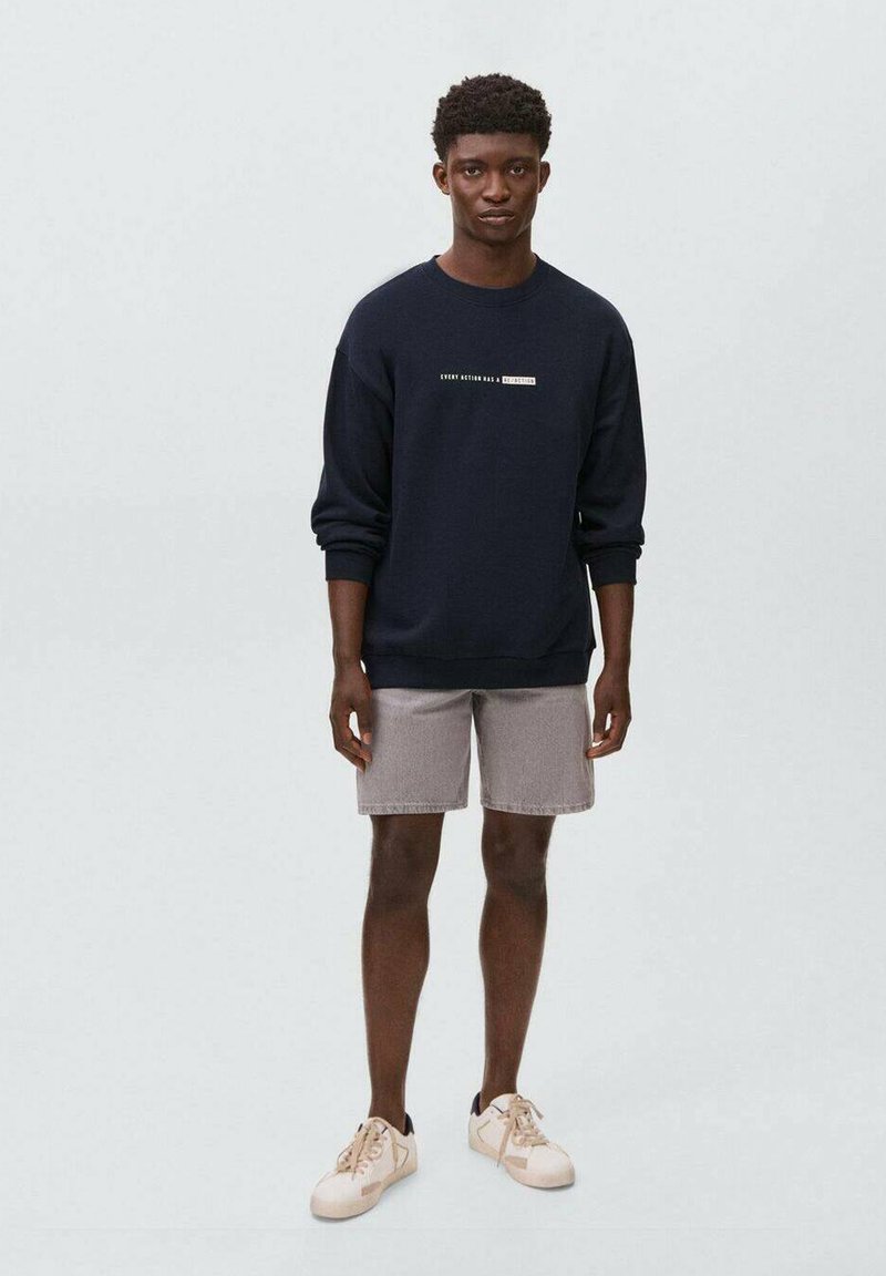 Felpa blu marina con testo, abbinata a pantaloni corti grigi chiari e sneaker beige. La felpa presenta un collo rotondo e polsini a costine.