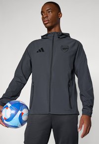 Mørkegrå hettejakke med glidelås, med Arsenal-logo og tre svarte striper. Glatt tekstur med elastiske mansjetter, sammen med en fargerik fotball.