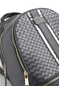 Černý batoh s tkanou texturou, hladkými panely, zlatými kovovými doplňky a předním logem s bílými akcentními pruhy.