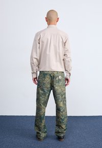 Camicia beige a maniche lunghe abbinata a pantaloni cargo mimetici verdi, con tasche e un design testurizzato. Modello girato di spalle su un tappeto blu.