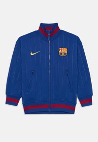 Veste de sport bleue avec des accents rouges, fermeture éclair à l'avant, poignets et ourlet côtelés. Présente un logo de club et le swoosh de Nike en jaune. Design à rayures.