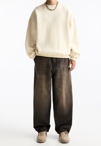 Beżowy, oversize sweatshirt z okrągłym dekoltem, w zestawie z luźnymi, ciemnoszarymi dżinsami. Stopy w beżowych sneakersach. Płaskie, teksturowane powierzchnie.