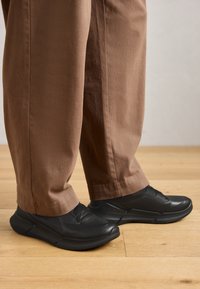 Person wearing loose brown pants and black slip-on sneakers standing on wooden floor.