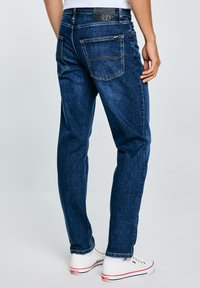 Jeans de mezclilla azul oscuro con diseño de pierna recta, que cuentan con costuras naranjas, cinco bolsillos y un parche con el logo en la cintura.
