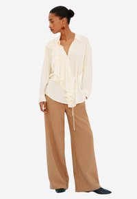 Blusa crema con frontale arricciato, collo alla francese, maniche lunghe, vita legata, abbinata a pantaloni marroni a gamba larga e scarpe nere slip-on.