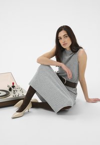 Femme aux longs cheveux foncés portant une robe grise sans manches, des collants marron et des talons beiges, assise par terre à côté d'un tourne-disque.