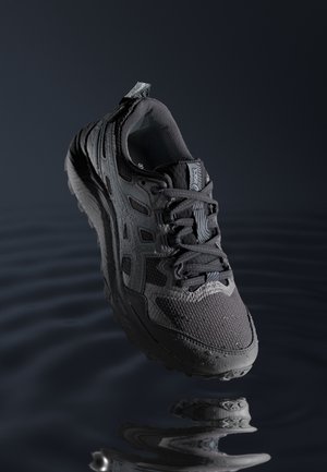 Dunkelgrauer ASICS-Laufschuh mit detaillierter Textur und Schnürsenkeln, schwebt über Wasser mit einer wellenförmigen Spiegelung darunter.