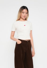Witte geribbelde short-sleeve t-shirt met rode hartborduursel; gecombineerd met bruine corduroy wijde broekspijpen. Eenvoudig, casual ontwerp.