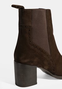 Bruine leren enkellaars met elastische zijkanten, decoratieve perforaties en een stevige zwarte houten hak. Beschikt over een trekhaak aan de achterkant.