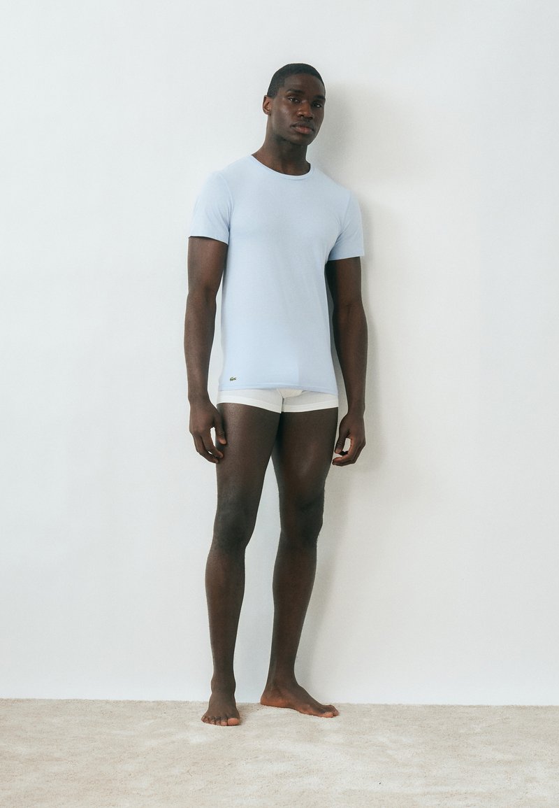 Jeune homme debout pieds nus contre un mur blanc, portant une chemise à manches courtes bleu clair et un short blanc.