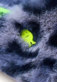 Tejido gris oscuro y esponjoso con un personaje de plástico amarillo sonriente parcialmente visible, destacando una textura suave y un diseño juguetón.