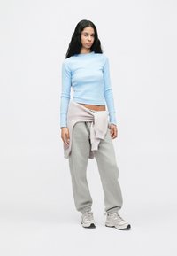 Νεαρή γυναίκα που στέκεται φορώντας ένα ανοιχτό μπλε πουκάμισο με μακριά μανίκια, γκρίζα φόρμα, απαλό ροζ φούτερ δεμένο στη  μέση, και μπεζ αθλητικά παπούτσια.