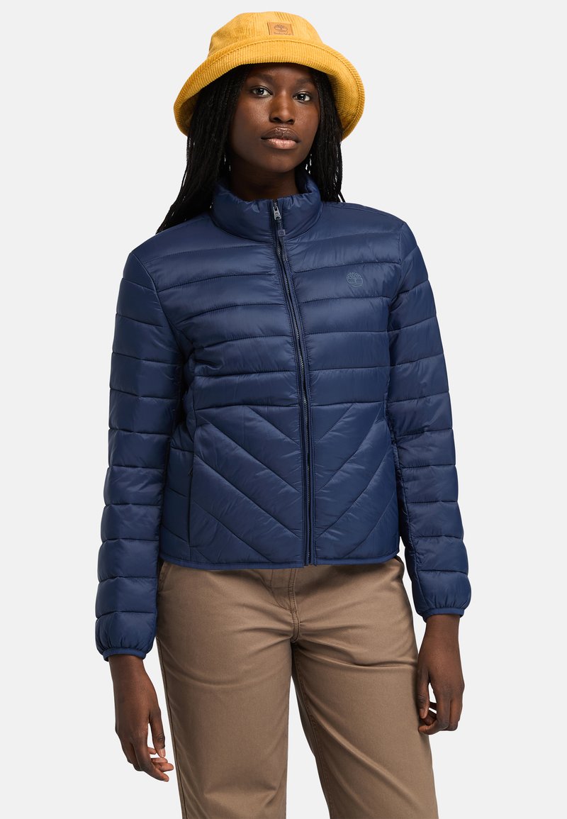 Giacca puffer blu navy con cuciture a zigzag, chiusura con zip e colletto alto. Modello con pantaloni beige e cappello a secchiello giallo.