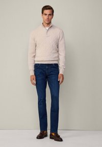 Lys beige kabelstrikket sweater med en kvart lynlås, parret med mørkeblå jeans og brune loafers. Ensfarvet baggrund, neutrale farvetoner.