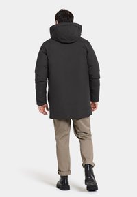 Giacca a vento nera con cappuccio, con finitura opaca e un taglio più lungo, abbinata a pantaloni khaki e stivaletti neri.