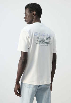 Hombre de piel oscura que lleva una camiseta blanca con un diseño tropical de un edificio y texto en la parte trasera, combinado con vaqueros de mezclilla azul claro.