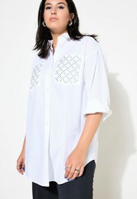 Camicia bianca con maniche arrotolate, caratterizzata da due tasche texturizzate adornate con borchie in metallo. Indossata con pantaloni a righe.