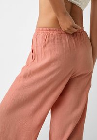 Personne ajustant la taille d'un pantalon loose, texturé, de couleur corail, avec un taille élastique et une poche latérale, portant un haut en tricot beige.