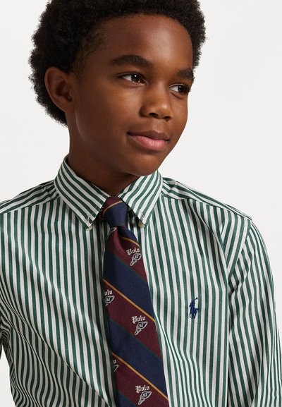 Zelena in bela črtasta srajca z ovratnikom na gumbe, kombinirana s temno modrim kratkim, ki ima maroon in rumene črte ter logotipe.