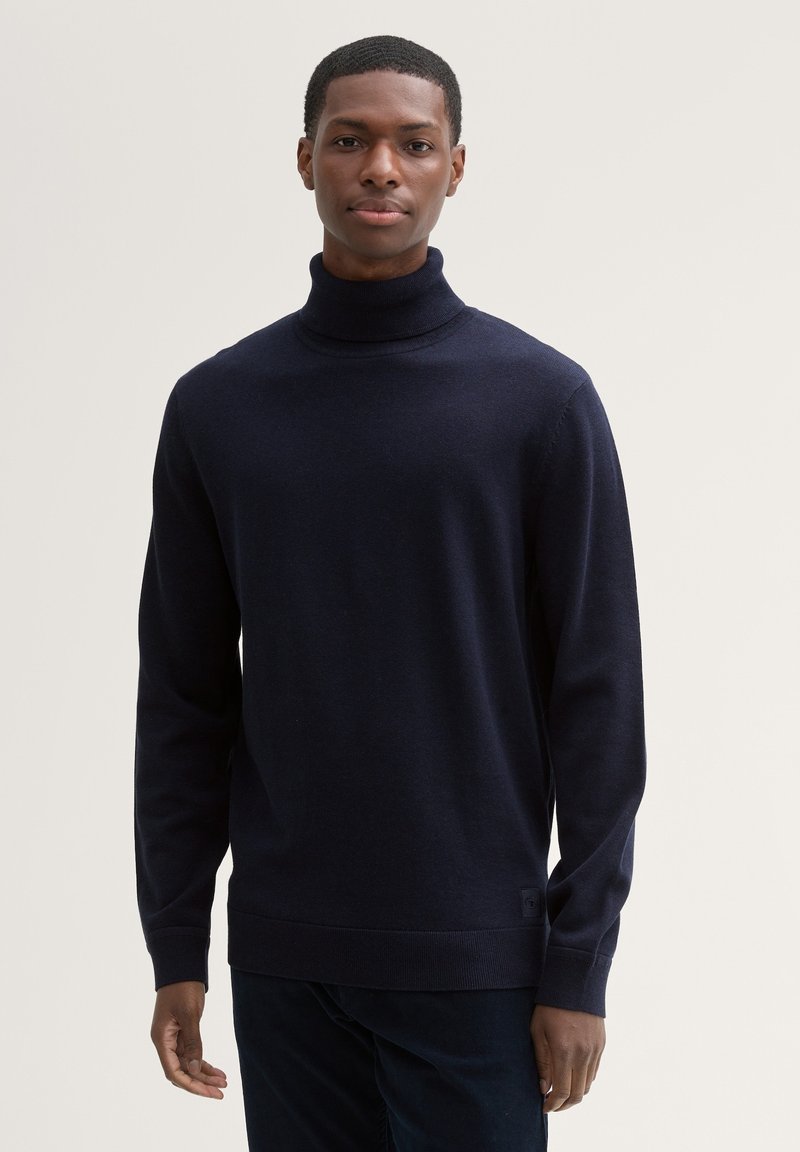 Granatowy sweter z wysokim kołnierzem wykonany z miękkiego materiału, z długimi rękawami i prążkowanym dołem. Dopasowany fason z minimalnym logo na dole.