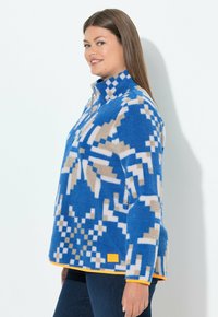 Niebieska bluza polarowa z geometrycznym wzorem w kolorze beżowym i białym. Wysoki kołnierz z półzamkiem, pomarańczowe wykończenie na dole i detal z logo.