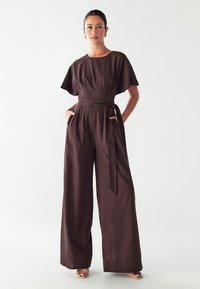 Jumpsuit de lino marrón con piernas anchas, mangas cortas y cintura atada, que cuenta con bolsillos laterales y una tela suave y texturizada.