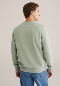 Sudadera de algodón verde con cuello redondo y puños acanalados, combinada con jeans azules. La tela parece suave con una textura lisa.