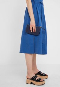 Jupe en lin bleu avec une fente sur le devant, maintenue par une ceinture, associée à des sandales à plateforme noires épaisses et à une petite pochette en cuir marine.
