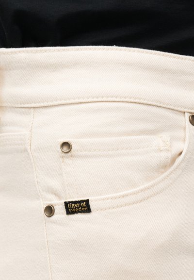 Κοντινό πλάνο σε τζιν μπεζ χρώματος με μπροστινή τσέπη και μαύρη ετικέτα της μάρκας "Tiger of Sweden" στην άκρη της τσέπης.