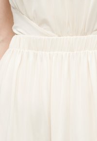 Κοντινή λήψη ενός ενδύματος εκρού με ελαστική μέση που μαζεύει και πλισέ λεπτομέρεια υφάσματος.