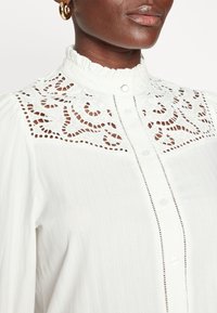 Gros plan d'une personne portant un chemisier blanc avec empiècement en dentelle, col à volants, fermeture boutonnée devant et boucle d'oreille dorée en forme d'anneau.