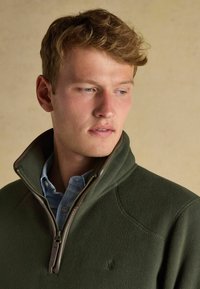 Giovane con i capelli castano chiaro, indossa un fleece verde con zip sopra una camicia a quadri blu, mentre guarda in basso e di lato.