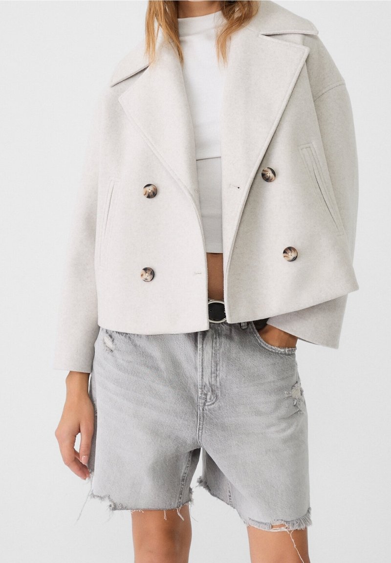 Personne portant un manteau court croisé beige clair sur un haut blanc avec un short en jean gris clair déchiré taille haute.