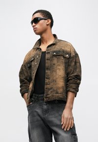 Smeđa traper jakna s blago izblijedjelom teksturom, s prednjim džepovima i srebrnim gumbima. Nošena preko crnog topa na bretele i sivih traperica. Crne sunčane naočale.