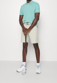 Světle modrá polokošile s límečkem, béžové šortky, bílé tenisky a golfová hůl držená vpředu, s stříbrnou sponou na pásku.