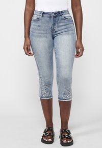 Femme portant un jean court bleu clair avec broderie florale près de l'ourlet et des sandales noires à boucles.