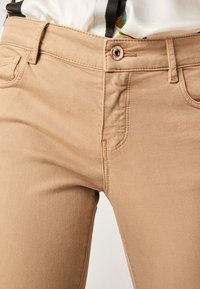 Pantalons beiges ajustés en tissu doux, avec une fermeture à bouton à l'avant, des passants de ceinture et un design classique à cinq poches.