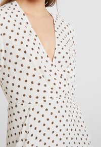 Vit klänning med djup V-halsringning, med ett mönster av bruna prickar. Slät tyg med en lätt glans, figurnära vid midjan.